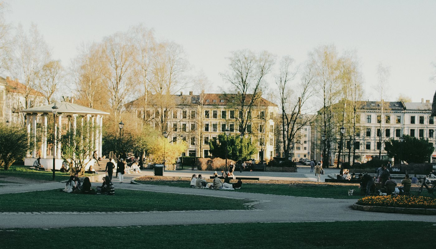 A gazebo and open lawn in a civic park, with people sitting on the grass.