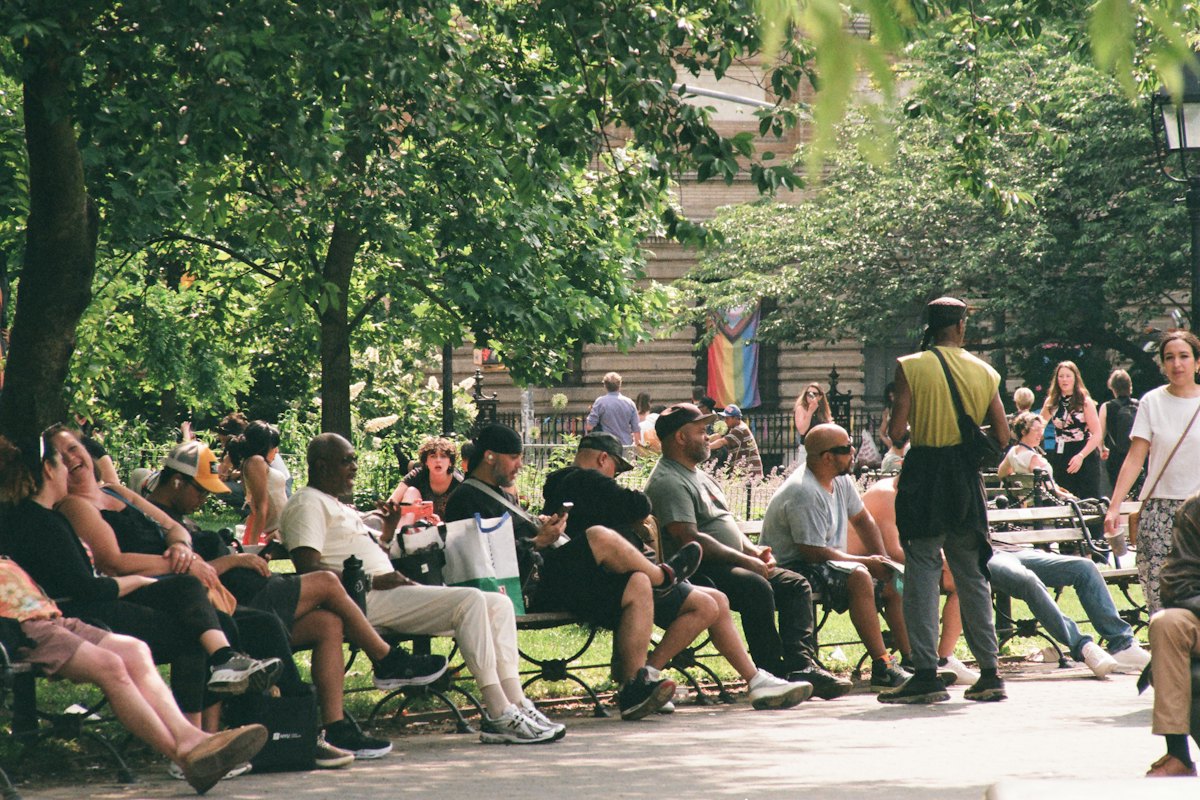 Residents sitting together on benches under shade trees in a busy park.